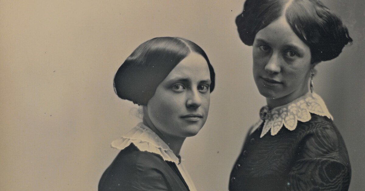 A black-and-white portrait photograph of two young White girls facing each another in period clothing and hairstyles.