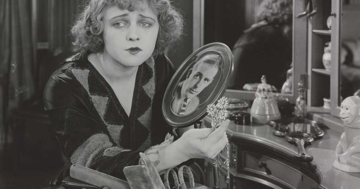 A black-and-white photograph of a White woman sitting at a vanity holding a brooch and a photograph of a White man as she gazes into the distance.